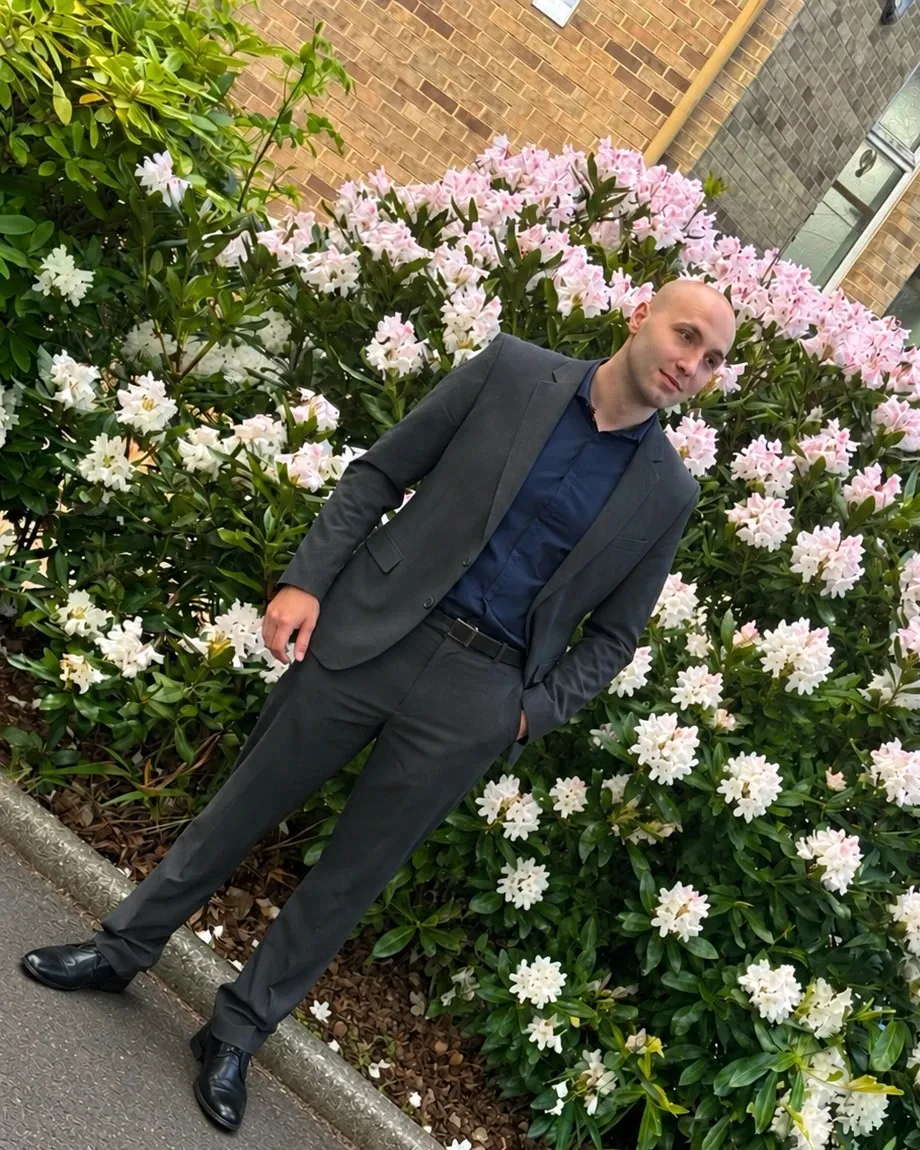 Sebastian Przyimka in a dark suit, standing in front of flowering bushes